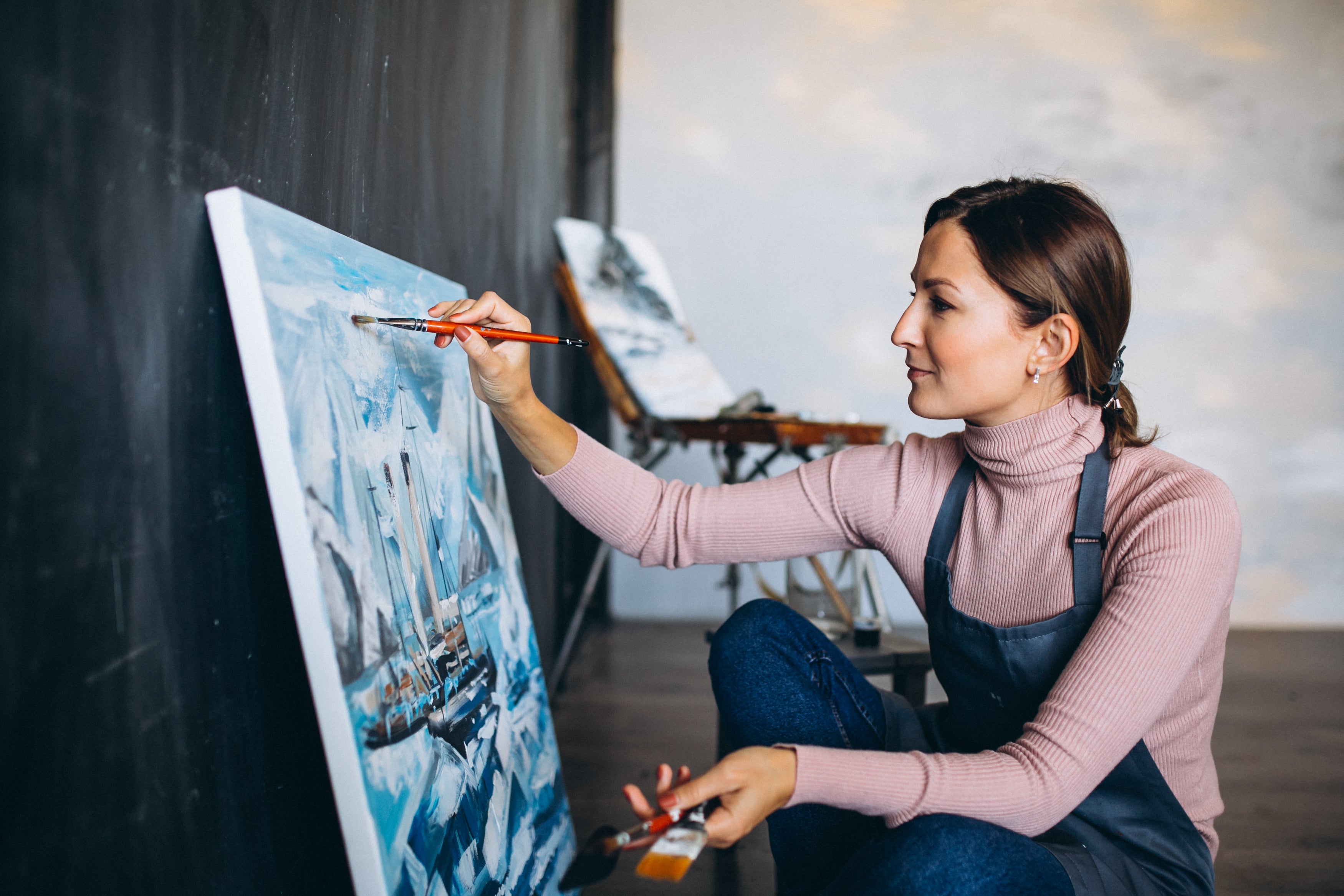 Woman painting a large canvas with a ship scene in an indoor setting.