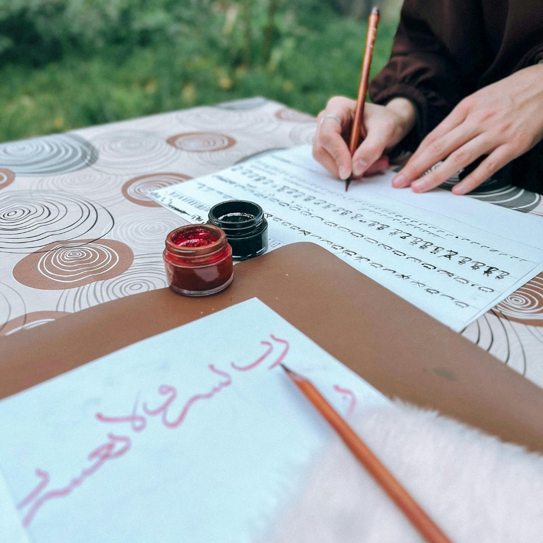 Person writing on paper with a pen, surrounded by ink containers outdoors.